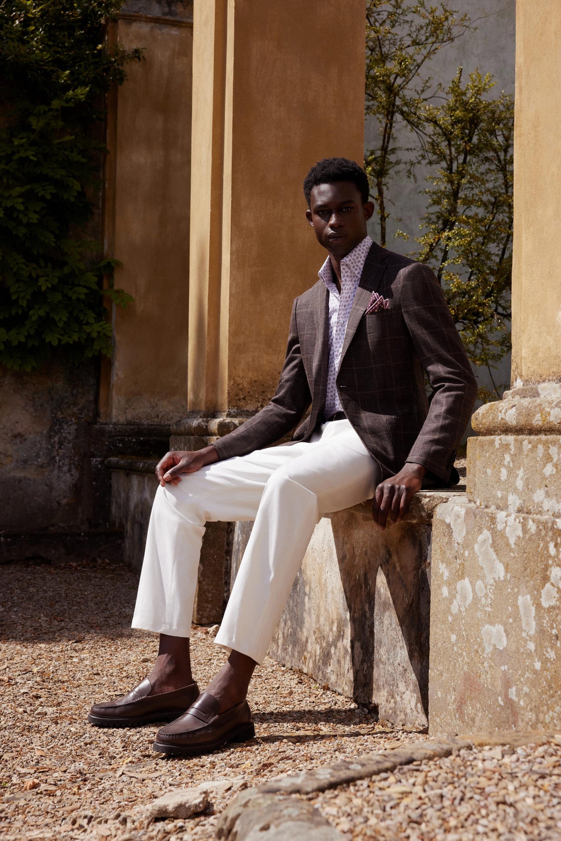 A man sitting on a stone bench, wearing a brown plaid sport coat with notch lapels, paired with a white shirt and beige trousers.